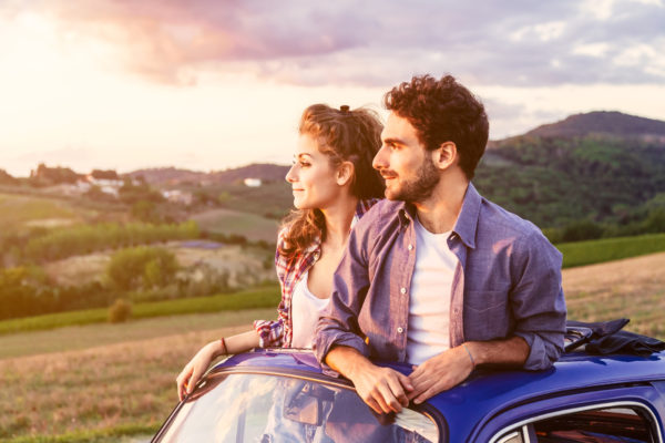 A loving couple, on a summer afternoon, watching sunset from the roof of an old car, around the classical landscape of Tuscany, vineyards and farmland. The two lovers come out from the car roof